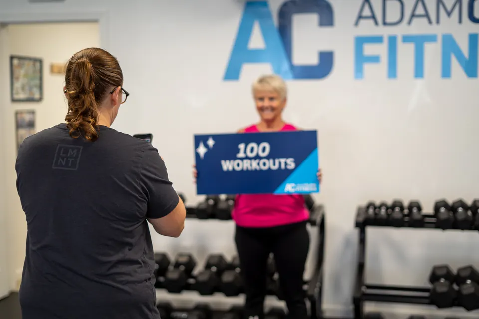 Adam Clark Fitness member holding a 100 workouts sign while a coach takes her photo