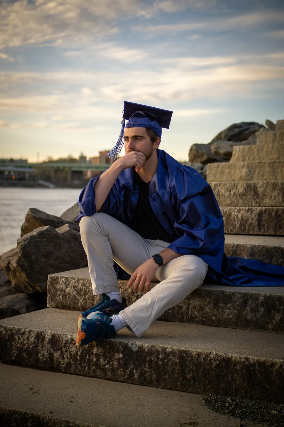Graduate in a blue gown sitting on stone steps by the water