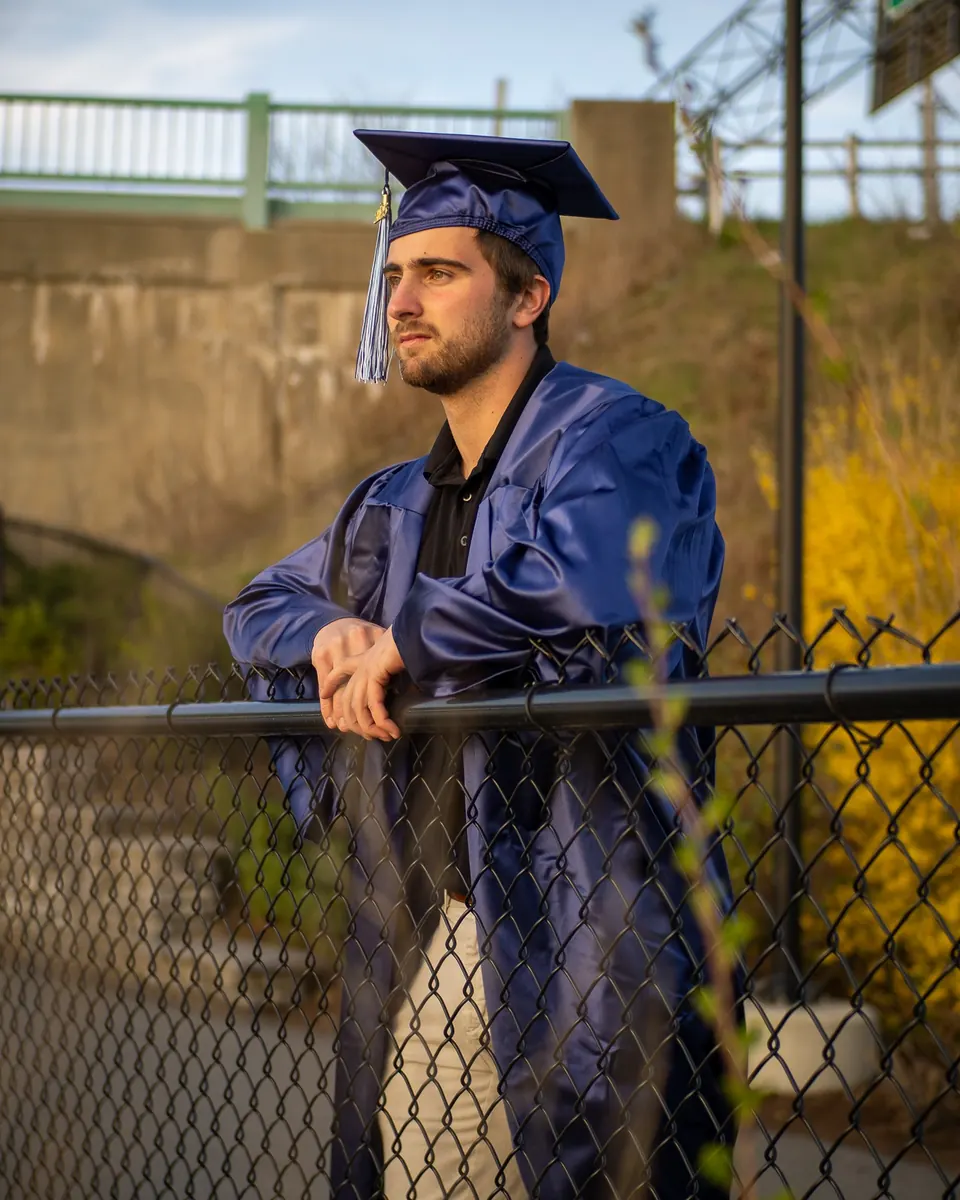 Graduate in a cap and gown leaning on a chain-link fence by fall trees