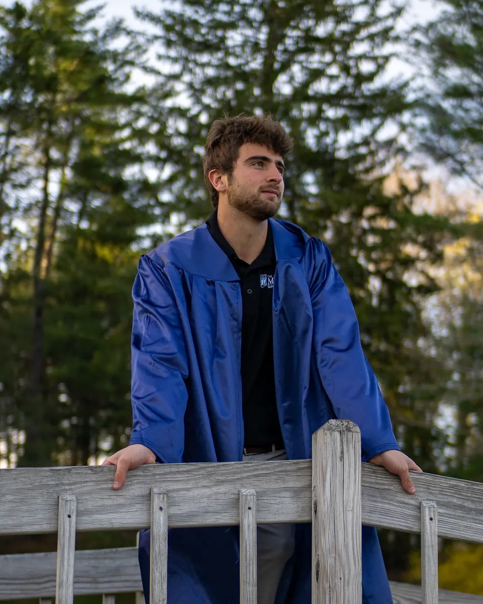Graduate in a blue gown leaning on a wooden rail in the woods